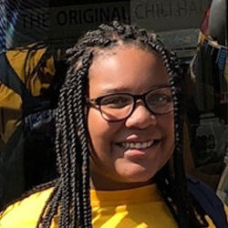 A young girl in a yellow shirt smiles in front of a building.