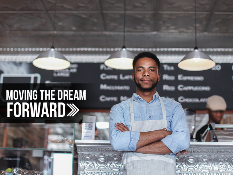 A man in an apron standing in front of a counter with the words moving the dream forward.