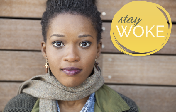 A woman with dreadlocks in front of a wooden wall with the words stay woke.