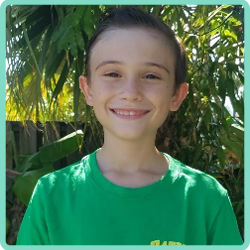 A boy in a green shirt is smiling in front of a palm tree.