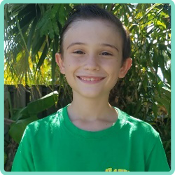 A boy in a green shirt is smiling in front of a palm tree.