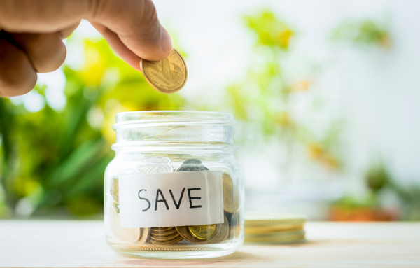 A hand putting a coin into a glass jar with the word save on it.