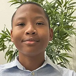 A boy in a blue shirt is smiling in front of a plant.