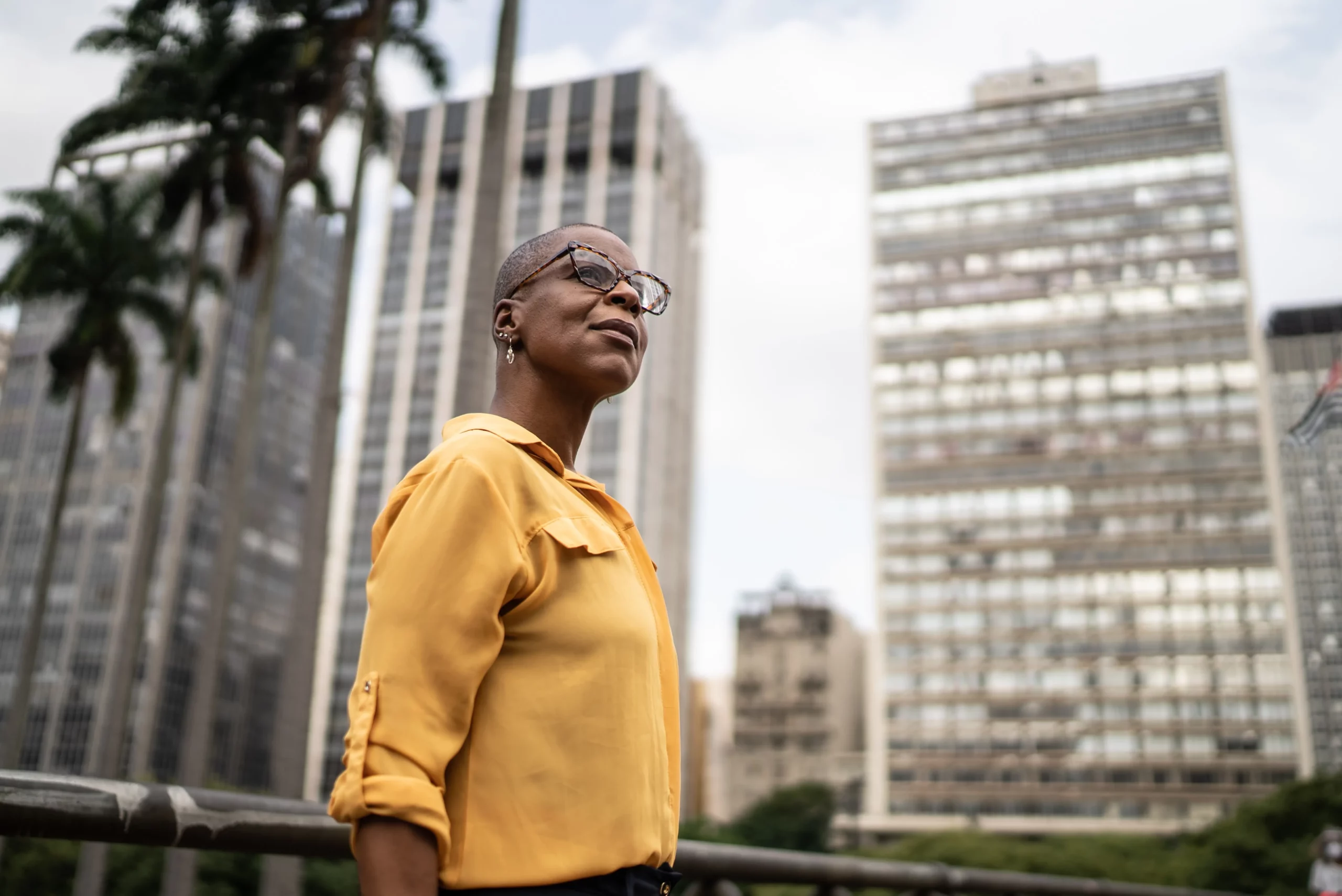 A black woman in a yellow shirt standing in front of tall buildings.