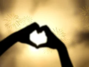 A silhouette of two hands making a heart shape with fireworks in the background.