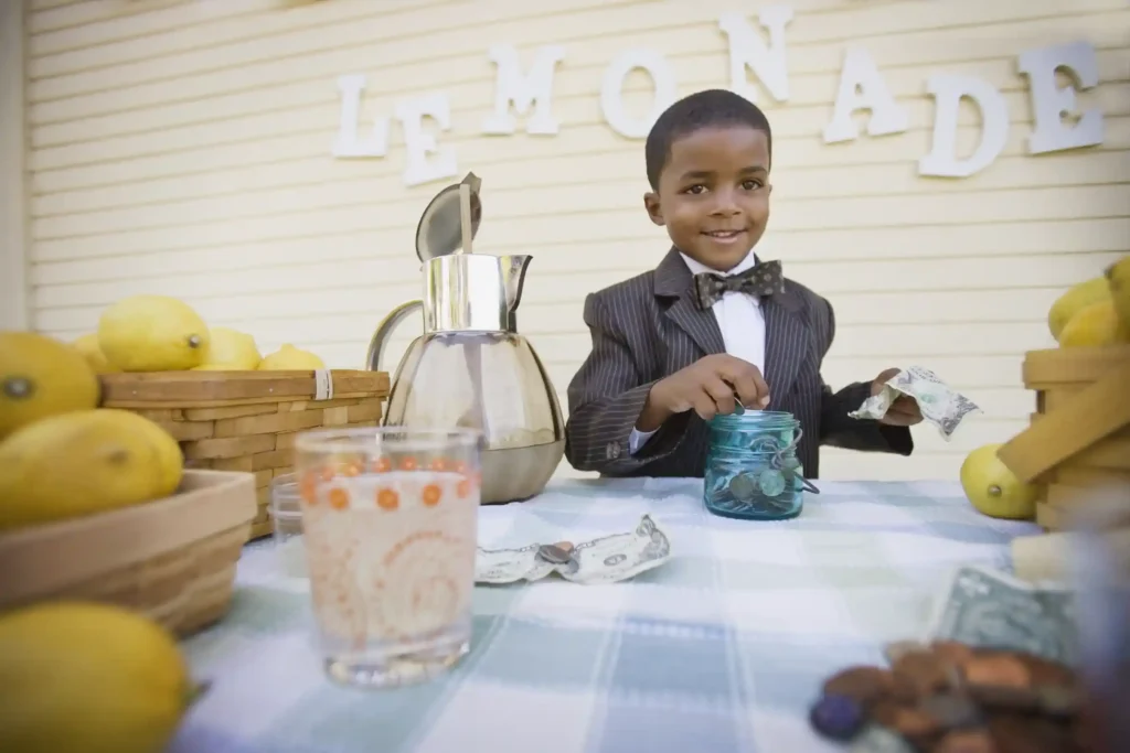 A young boy in a tuxedo sitting at a lemonade stand, learning about entrepreneurialism and experiencing the potential for generational wealth.