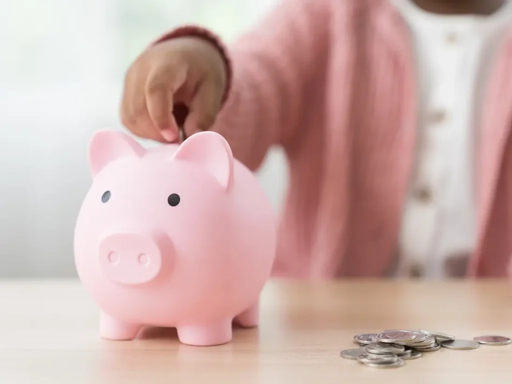 A child is enthusiastically depositing coins into a pink piggy bank, fostering the concept of generational wealth.