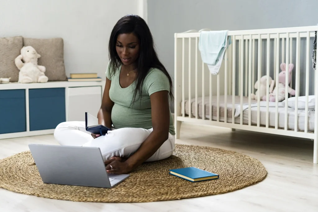 A pregnant woman sitting on the floor with a laptop in front of a crib.