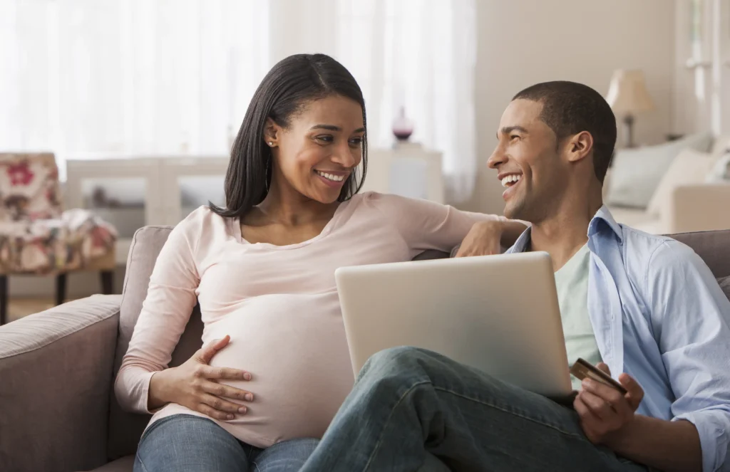 A pregnant man and woman sitting on a couch using a laptop.