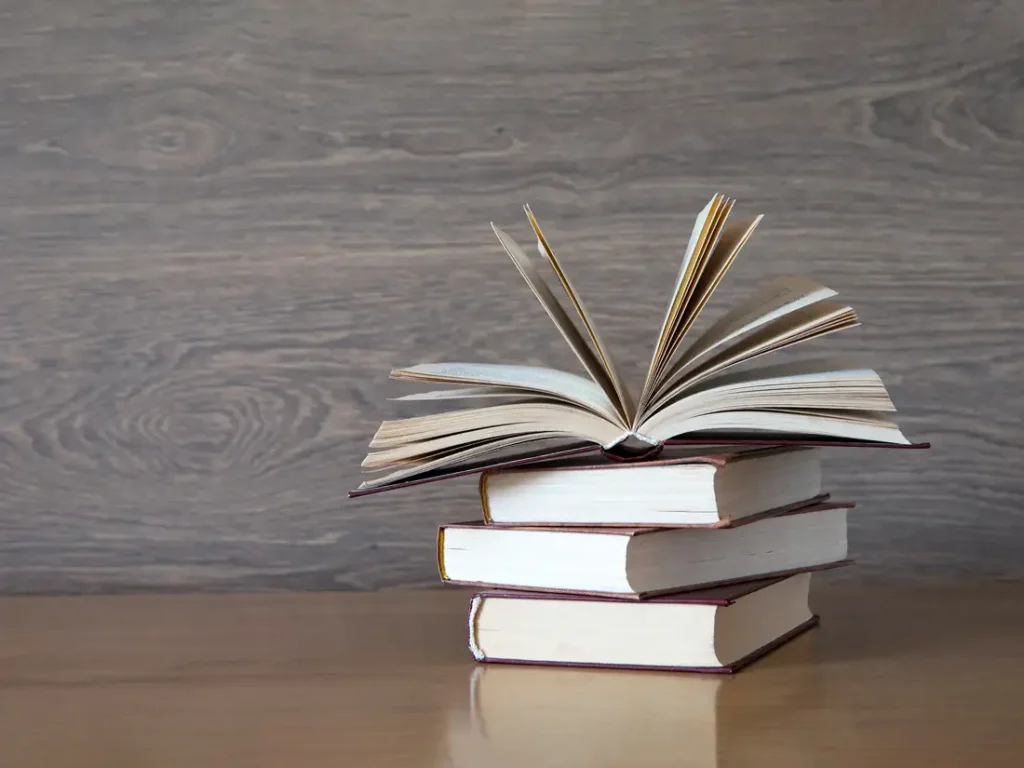 A stack of hardcover books with the top one opened on a wooden table and background.