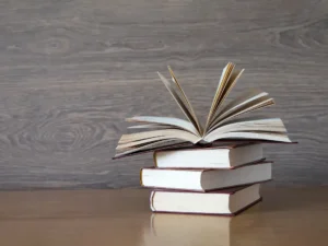 A stack of hardcover books with the top one opened on a wooden table and background.