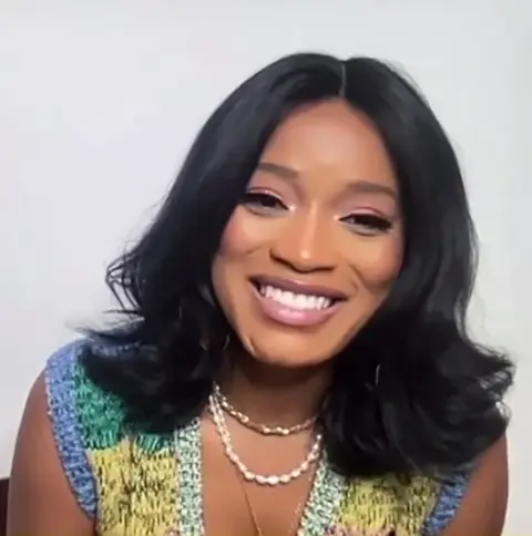 A woman with shoulder-length black hair smiling at the camera, wearing a colorful top and a pearl necklace.