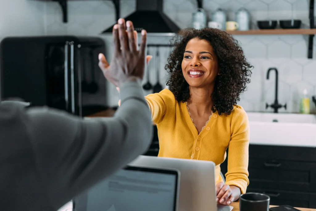 A woman in a yellow cardigan, engaged in purposeful purchasing, smiling and high-fiving someone across from her in a kitchen setting with a laptop in the foreground.