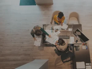 Top-down view of three people at a desk, including an intern, collaborating over documents and a laptop in a modern office setting.