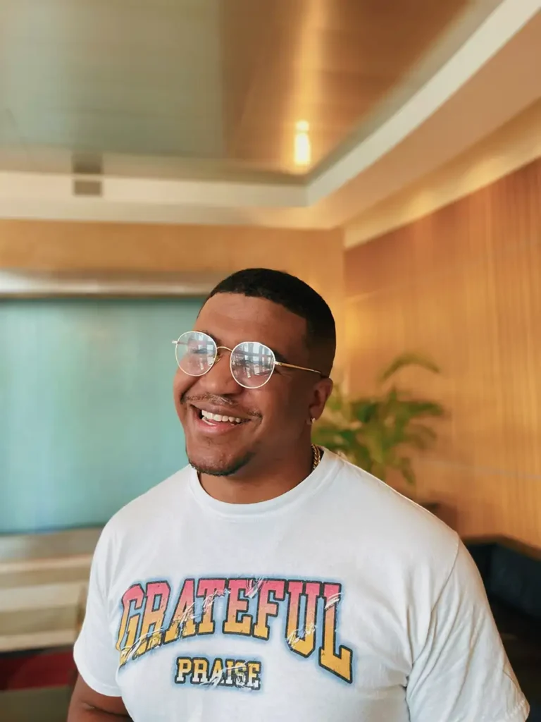 A smiling man wearing a "grateful praise" t-shirt and round glasses, indoors with a wooden wall background.