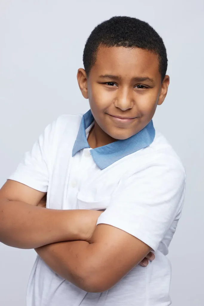 Confident boy with arms crossed smiling against a gray background.