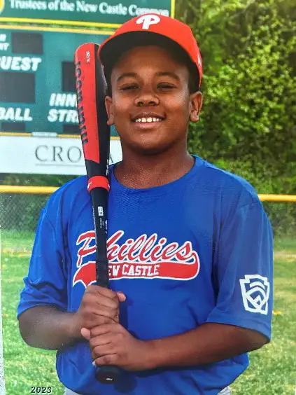 Young baseball player in a phillies jersey holding a bat, smiling on the field.
