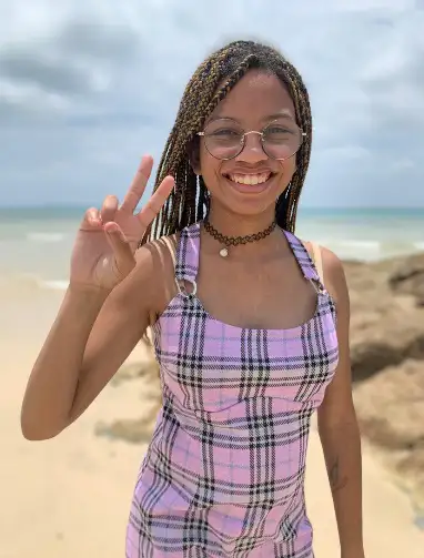 Young woman with braided hair and glasses making a peace sign on the beach.