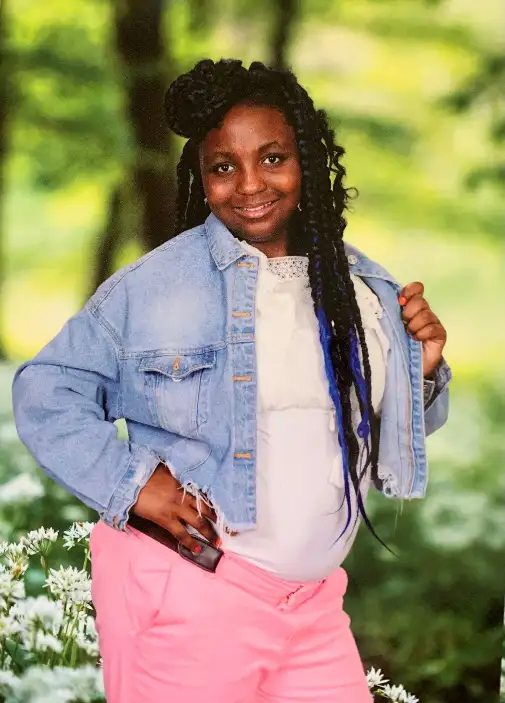 Young girl posing with a hand on her hip, wearing a denim jacket and pink pants, with braided hair, against a backdrop of greenery.