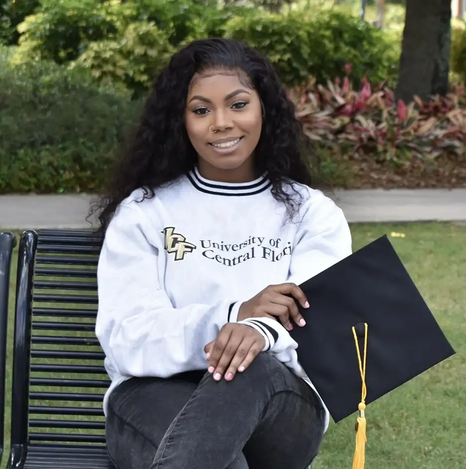 Young woman in a University of Central Florida sweatshirt sitting on a park bench holding a graduation cap.