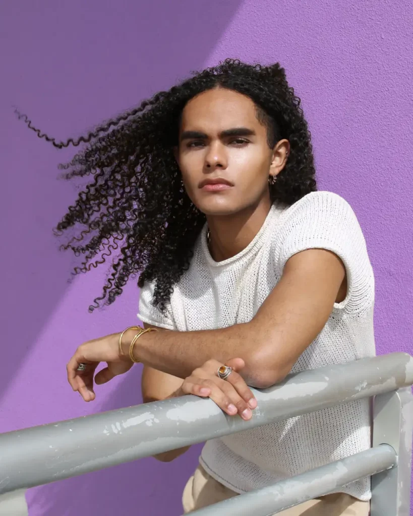 Young man with curly hair leaning on a railing against a purple wall, wearing a white top.