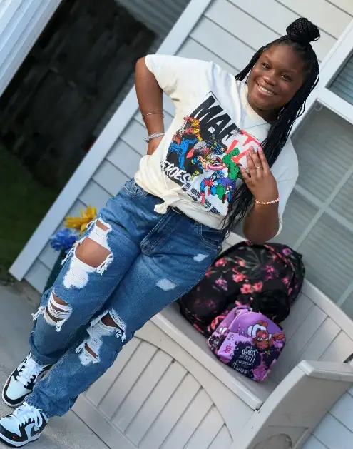A person smiling and posing outdoors, dressed in a graphic tee, ripped jeans, and sneakers, with a backpack on the steps behind.