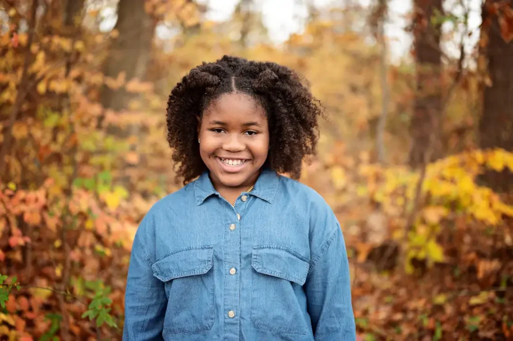 A cheerful young girl in a denim shirt stands before an autumnal forest backdrop.