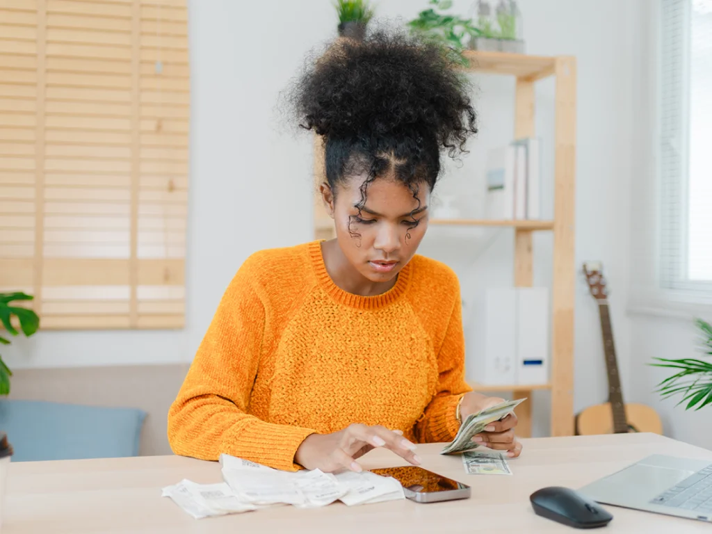 A young woman with hair styled in a high bun, wearing an orange sweater, appears to be managing her finances. She is sitting at a table with a laptop, a smartphone, cash, and receipts. The setting seems to be a casual living space, with a bookshelf and a guitar visible in the background.