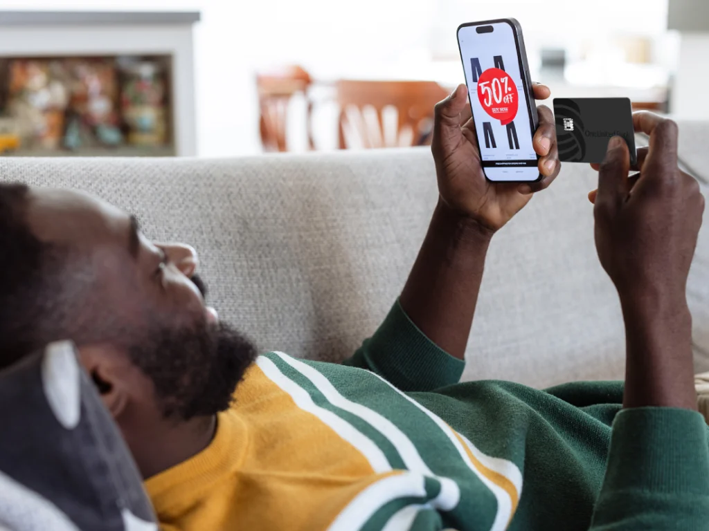 A man with a beard lies comfortably on a light gray couch, holding a smartphone in his right hand and a black credit card in his left. The phone displays a shopping website with a prominent red circle announcing "50% off" over an image of black pants. The man is looking at the phone with a slight smile, suggesting he's engaged in online shopping. He's wearing a dark green long-sleeved shirt over a yellow and white striped shirt. The background is slightly blurred but shows a living room setting with a white fireplace and hints of other furniture. The lighting suggests a bright, indoor environment.