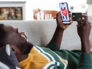 A man with a beard lies comfortably on a light gray couch, holding a smartphone in his right hand and a black credit card in his left. The phone displays a shopping website with a prominent red circle announcing "50% off" over an image of black pants. The man is looking at the phone with a slight smile, suggesting he's engaged in online shopping. He's wearing a dark green long-sleeved shirt over a yellow and white striped shirt. The background is slightly blurred but shows a living room setting with a white fireplace and hints of other furniture. The lighting suggests a bright, indoor environment.