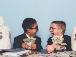 Two young boys in suits and glasses excitedly hold cash, surrounded by money bags, coins, a piggy bank, and a calculator on a table.
