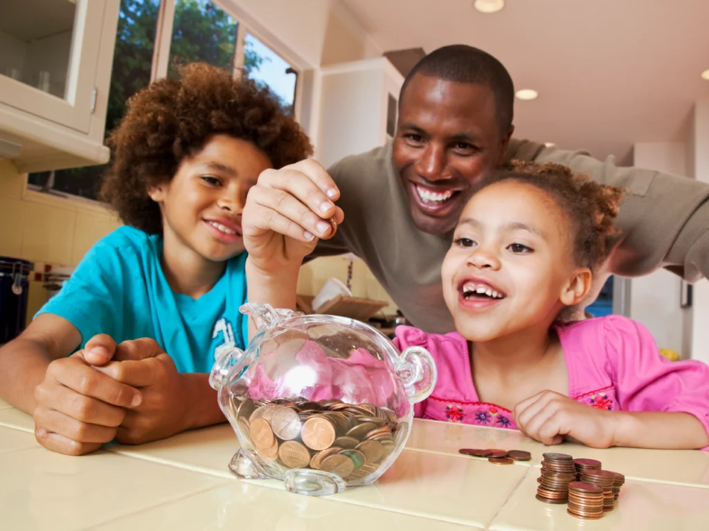 An adult and two children smile as they put coins into a transparent piggy bank on a kitchen counter, with stacks of coins nearby.