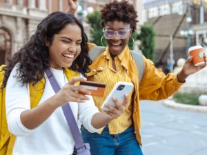 Two young women smile outdoors; one holds a smartphone and a credit card, while the other holds a coffee cup and raises her arm in excitement.