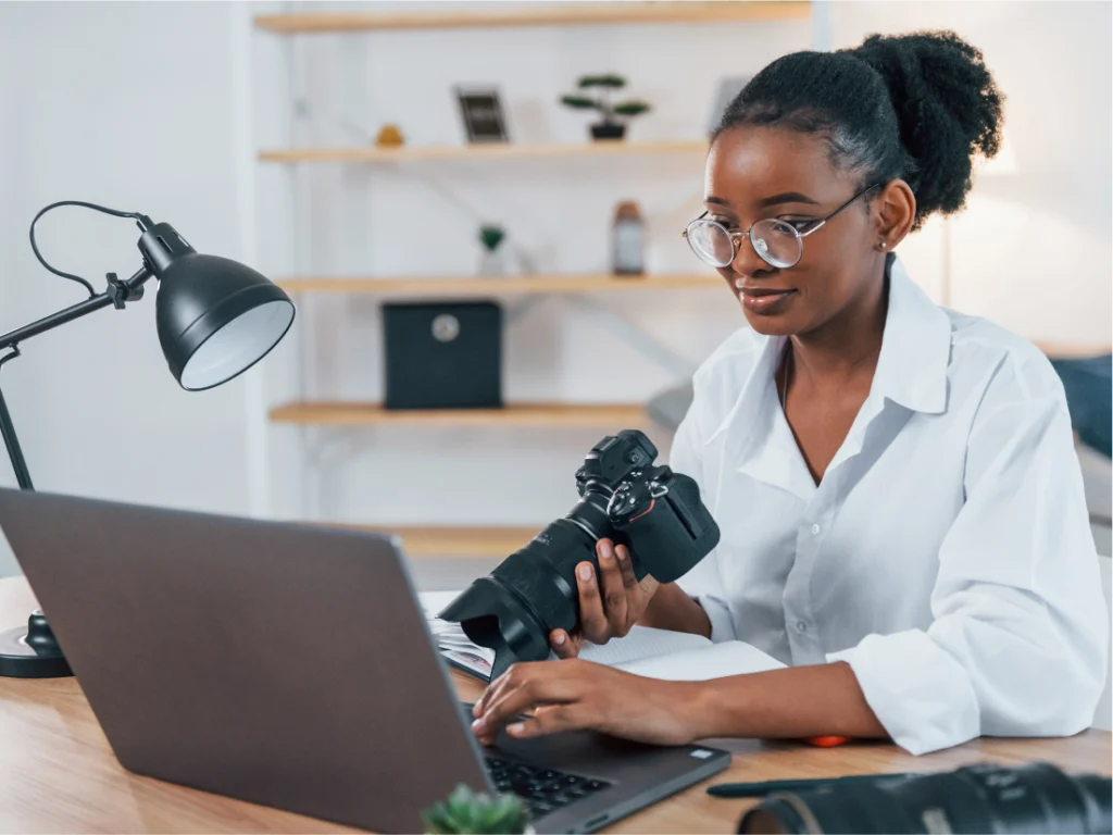 A woman wearing glasses sits at a desk, balancing gig economy work—holding a camera in one hand and using a laptop with the other. A lamp and camera lens are also on the desk.