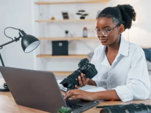 A woman wearing glasses sits at a desk, balancing gig economy work—holding a camera in one hand and using a laptop with the other. A lamp and camera lens are also on the desk.