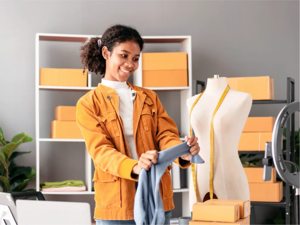 A person in a yellow jacket, working in the gig economy, holds a piece of fabric in front of a dress form with a measuring tape, surrounded by boxes and shelves in a busy workspace.