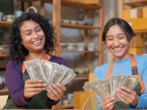 Two women in aprons smile and hold multiple $100 bills each, showing off the rewards of their side hustle indoors, with shelves of pottery and boxes in the background.