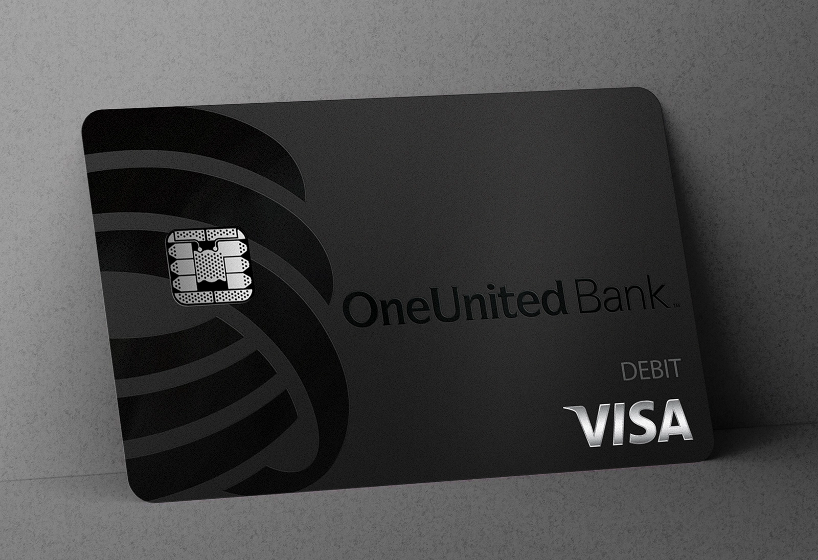 A black OneUnited Bank Visa debit card with a chip is propped against a gray surface.