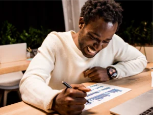 A man in a white sweater sits at a desk, smiling while analyzing and marking a colorful bar chart on a piece of paper with a pen.