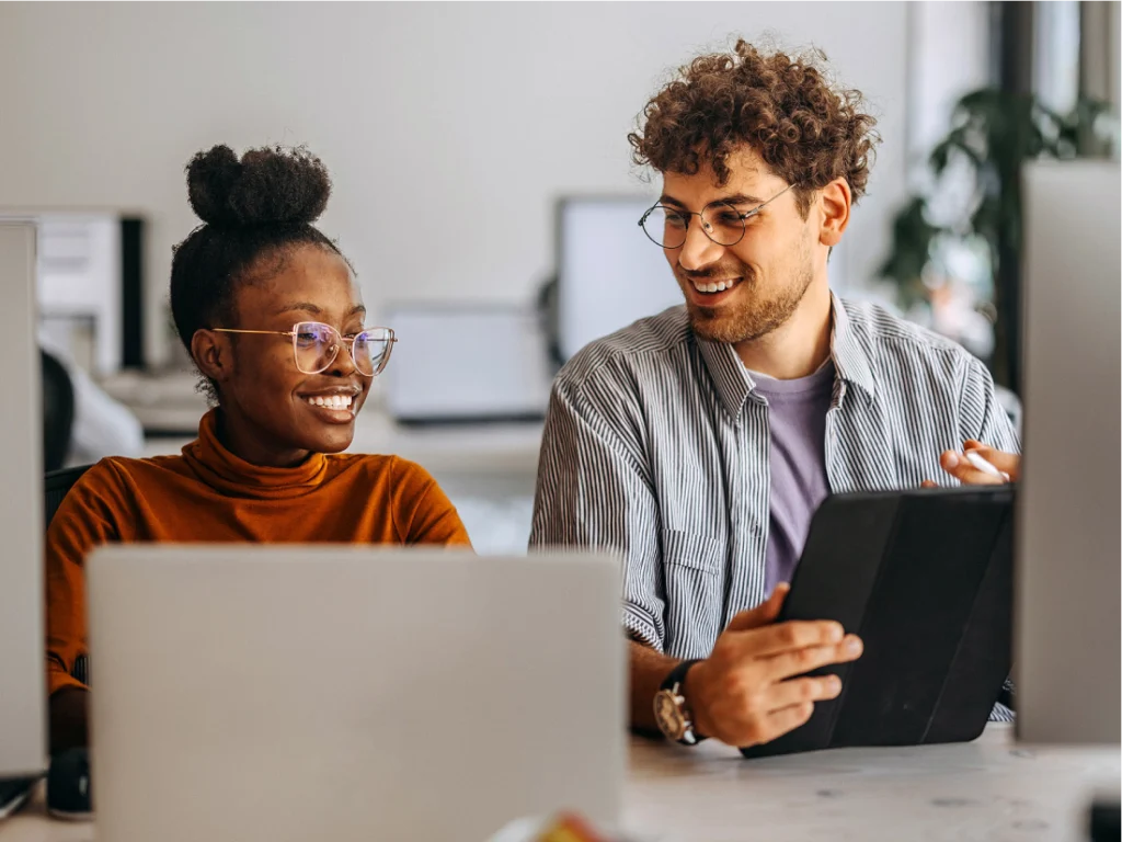 Two people sit at a desk with laptops, smiling and talking; one holds a tablet while the other looks at them.