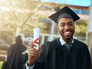 A graduate in a cap and gown smiles while holding a diploma outdoors on a sunny day. Other graduates and trees are visible in the background.