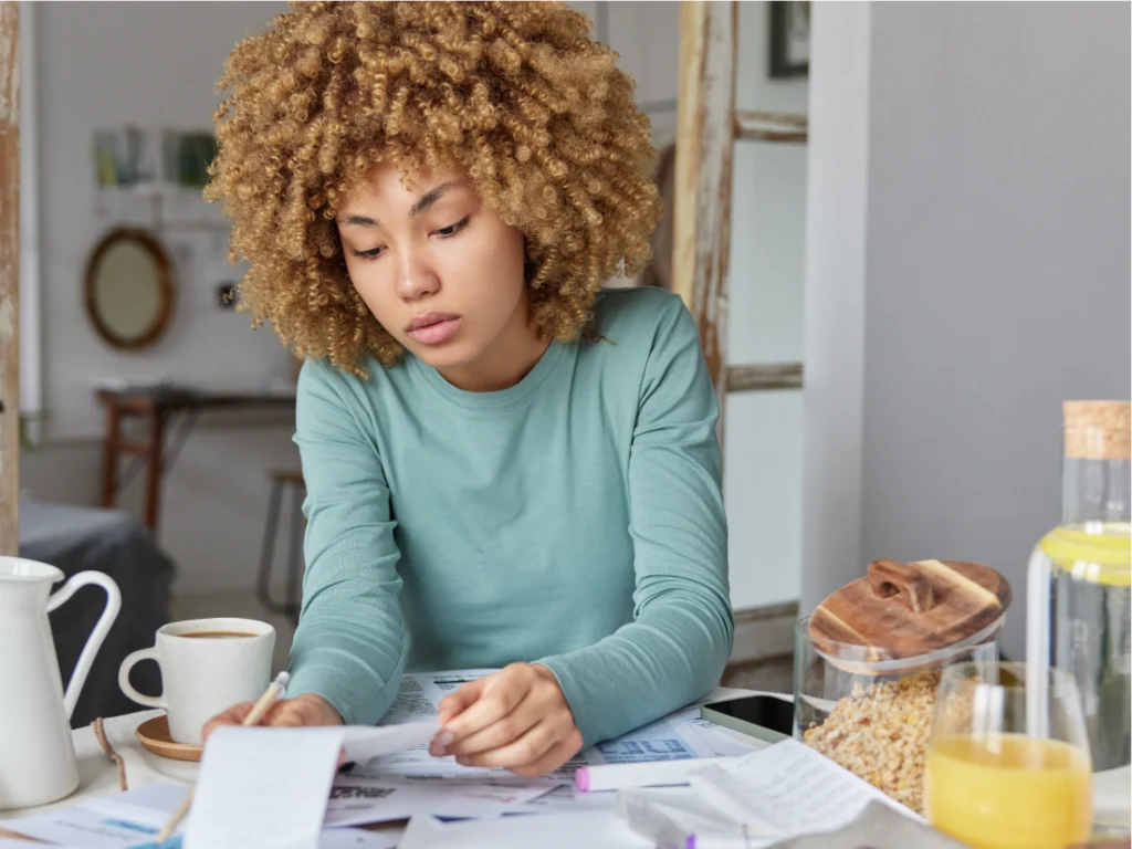 A woman with curly hair reviews papers at a table with a cup of coffee, orange juice, cereal, and documents spread out in front of her.