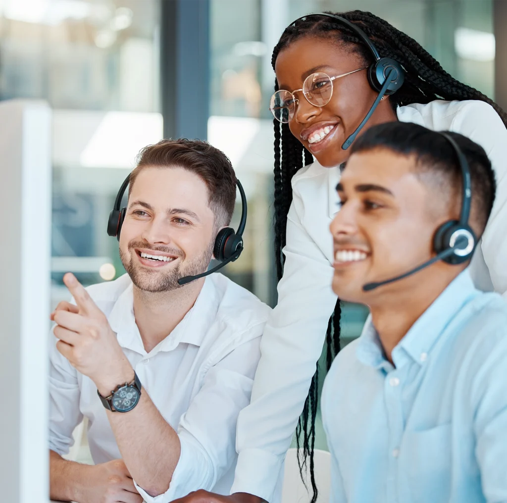 Three customer service representatives wearing headsets work together at a computer, smiling and collaborating in a modern office.