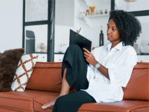 A woman sits on a brown sofa with her legs folded, writing about her Financial Detox plan on a clipboard in a modern living room with a kitchen in the background.