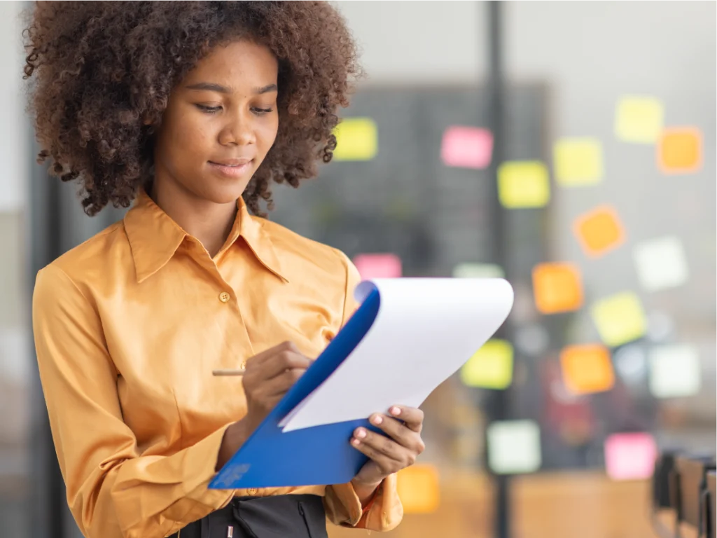 A person in a yellow shirt writes on a clipboard, standing in front of a window covered with colorful sticky notes, planning their Financial Detox.
