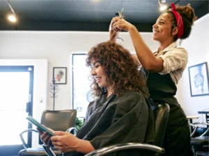 A hair stylist cuts the curly hair of a seated client, who is reading a tablet in a modern, well-lit salon.