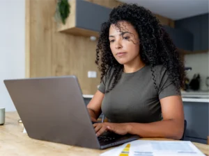 A woman with curly hair works on a laptop at a kitchen table with documents and a pen nearby.