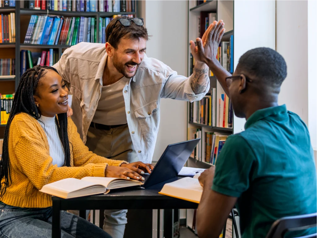 Three people in a library sit at a table with books and a laptop; one person stands, smiling and giving a high five to another seated person.