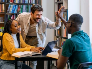 Three people in a library sit at a table with books and a laptop; one person stands, smiling and giving a high five to another seated person.