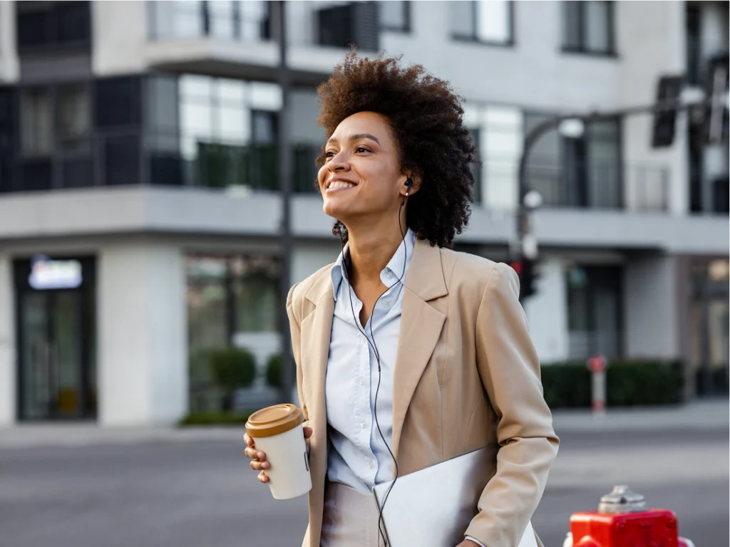 A woman in a beige suit holds a coffee cup and laptop, smiling while walking outside near modern buildings, enjoying a rest reset as she starts her day.